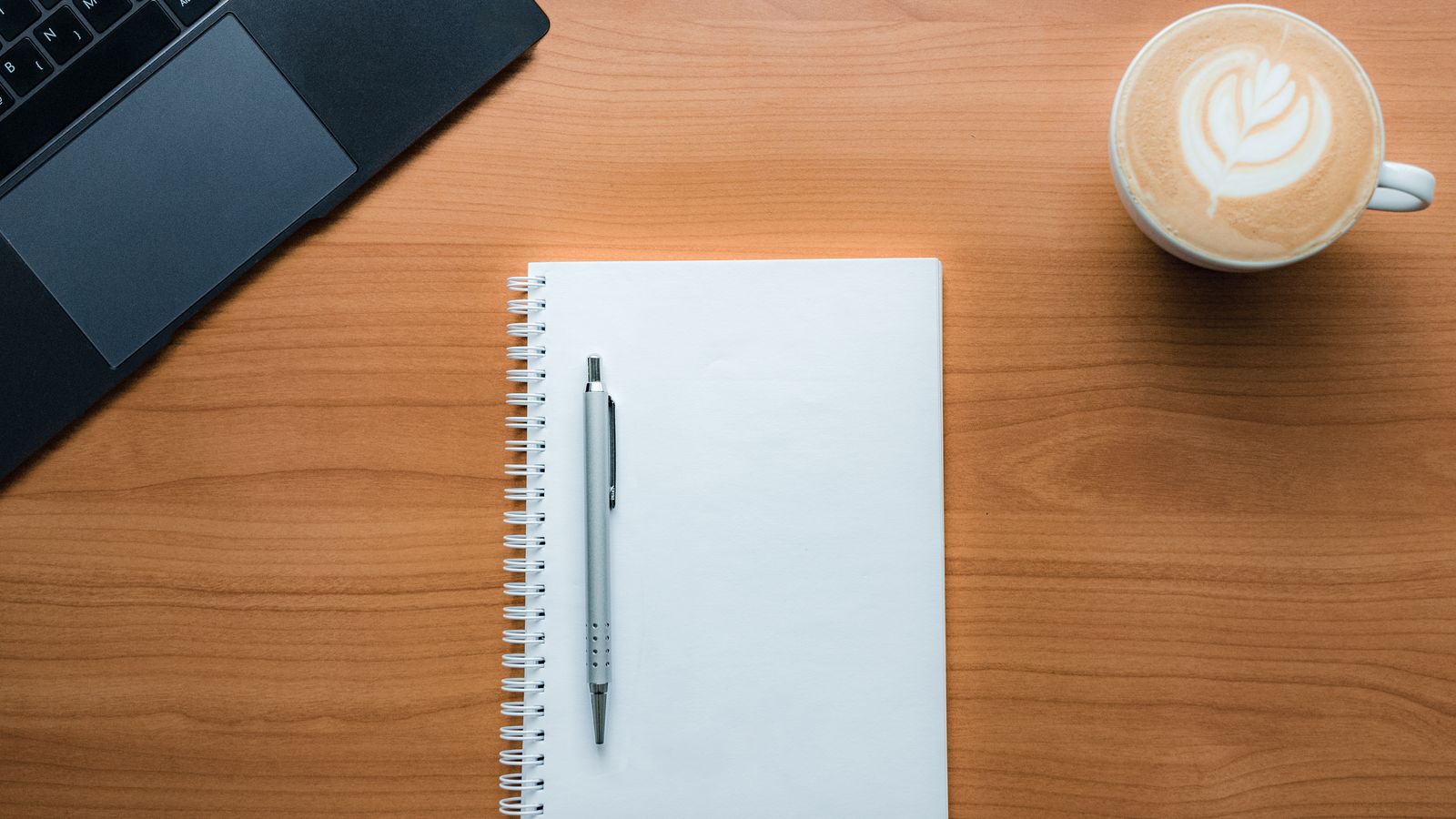 A blank notebook, pen, laptop and a cup of coffee on a wooden desk